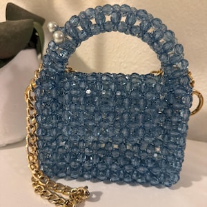 Puede incluir: Un pequeño bolso de mano cuadrado hecho de cuentas azul claro. El bolso tiene una correa de cadena dorada y un asa a juego. Dos cuentas de perlas blancas están unidas al asa. El bolso está sobre una superficie blanca.