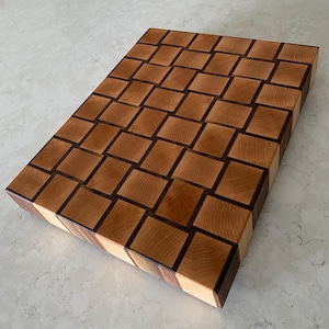May include: A rectangular end-grain cutting board with a checkerboard pattern. The board is made of light and dark brown wood blocks, creating a visually appealing design. The wood grain is visible, adding to the natural aesthetic.