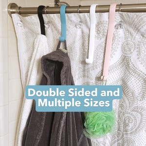 May include: Shower curtain hooks in black, blue, white, and pink hang from a metal rod. A white towel and a brown hooded robe hang from the hooks. A green loofah is also hanging. The text "Double Sided and Multiple Sizes" is in a blue box.
