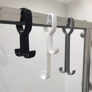 May include: Three shower door hooks in black, white, and grey hang from a metal bar. Each hook has a curved top and a double hook design at the bottom, suitable for hanging towels or other bathroom accessories.