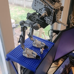 May include: A gray plastic building block model of a bipedal walker vehicle with a cockpit and two legs, standing on a blue rectangular base plate. A small gray and white plastic minifigure stands on the base plate next to the walker vehicle.