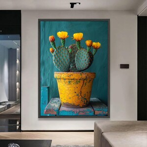 May include: A framed canvas print depicting a cactus in a yellow, weathered pot. The cactus has green pads and is decorated with yellow and orange flowers. The background is a textured teal colour, and the pot sits on a blue wooden surface.