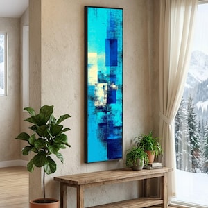 May include: Vertical abstract art piece with a black frame. The artwork features a blend of blue, teal, and white rectangular shapes. The piece is displayed on a beige wall, with a wooden bench and plants in front of it. A window with a snowy mountain view is visible.