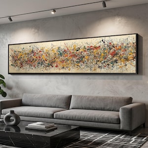 Puede incluir: Pintura expresionista abstracta enmarcada en negro. La obra presenta un fondo beige con salpicaduras de pintura roja, amarilla, negra y azul. El lienzo rectangular largo está montado sobre un sofá gris.