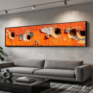 May include: A rectangular framed artwork featuring a close-up of a weathered orange surface with several dark holes. Patches of gray and white material are visible, adding texture. The artwork is displayed above a gray sofa in a modern living room setting.