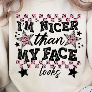 Könnte beinhalten: Cremefarbenes Sweatshirt mit dem Text "I'M NICER than MY FACE looks" in Schwarz und rosa Leopardenmuster. Das Design umfasst Sterne und einen Leopardenmuster-Rand. Das Sweatshirt ist ein lässiger, bequemer Stil.