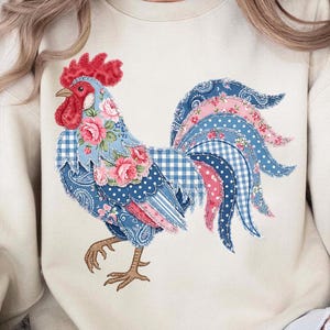 May include: Cream-colored sweatshirt featuring a colorful rooster design. The rooster is adorned with floral, paisley, and gingham patterns in shades of blue, pink, and red. The sweatshirt has long sleeves and a crew neck.