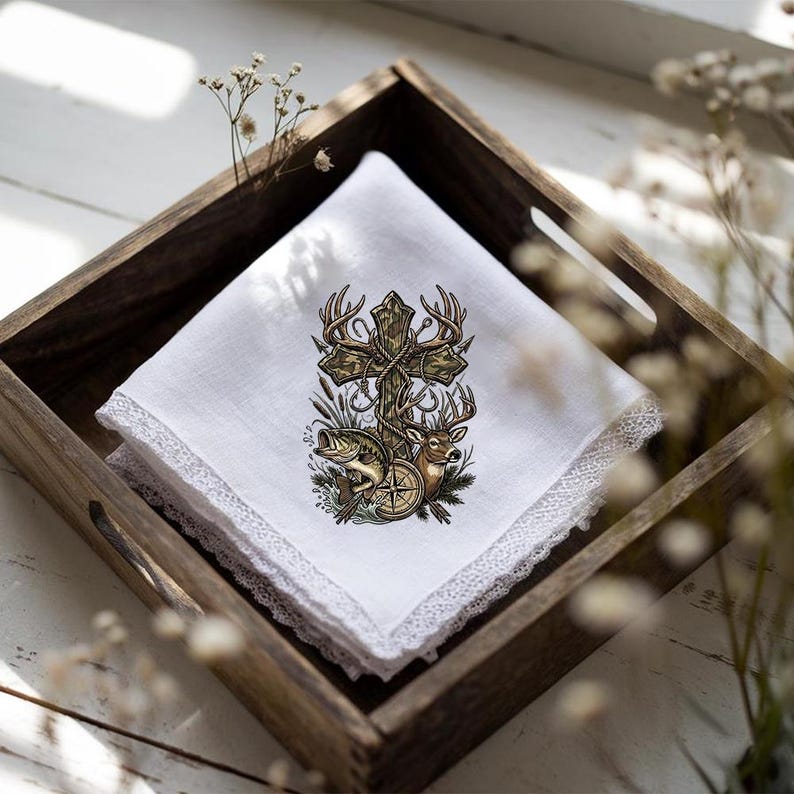 May include: White handkerchief with a decorative cross design featuring deer antlers, a fish, and a compass. The handkerchief has a delicate lace trim and is displayed in a wooden tray.