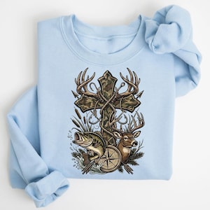 May include: Light blue sweatshirt featuring a graphic of a wooden cross with camouflage pattern, deer antlers, and fishing hooks. A deer, a compass, and a fish are also depicted. The design has a hunting and fishing theme.