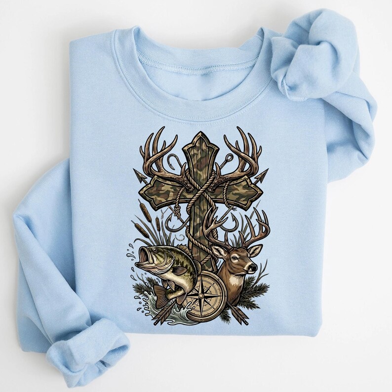 May include: Light blue sweatshirt featuring a graphic of a wooden cross with camouflage pattern, deer antlers, and fishing hooks. A deer, a compass, and a fish are also depicted. The design has a hunting and fishing theme.