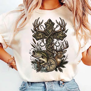 May include: Cream-colored t-shirt featuring a detailed graphic. The design includes a cross with deer antlers, a compass, a deer head, a fish, and fishing hooks. The cross has a camouflage pattern, and the overall theme is hunting and outdoors.