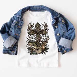 May include: White t-shirt featuring a cross design with deer antlers, a compass, a fish, and a deer. The cross is decorated with a camouflage pattern and rope. The shirt is paired with a blue denim jacket.