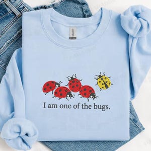 May include: Pale blue sweatshirt with an embroidered design of ladybirds. Four are red with black spots, and one is yellow. The text "I am one of the bugs" is below the ladybirds. The sweatshirt is made of a soft material.