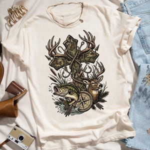May include: Cream-colored t-shirt featuring a graphic design with a cross, deer antlers, a compass, a fish, and a deer. The cross has a camouflage pattern. The design incorporates fishing hooks and arrows.