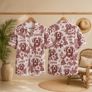 Puede incluir: Dos camisas de manga corta con botones que presentan un estampado repetitivo en color granate de caras de perro, flores de hibisco tropicales y hojas de palma sobre un fondo blanco. La palabra COOPER está impresa en granate sobre cada perro.