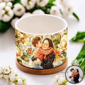 May include: A cylindrical ceramic planter with a white interior and a wooden base. The exterior features a colourful mosaic-style print of a person hugging a child, surrounded by detailed illustrations of white and orange magnolia flowers.