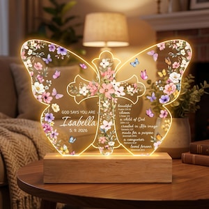 May include: A butterfly-shaped LED night light with a cross design. The clear acrylic light features floral and butterfly accents, the name "Isabella", and inspirational text. It rests on a wooden base and is illuminated with a warm glow.