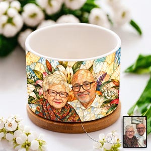 May include: A cylindrical ceramic planter featuring a colorful stained-glass style illustration of an older couple wearing glasses. The design includes vibrant floral patterns in the background and the planter rests on a circular wooden base.