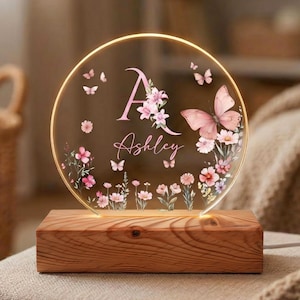 Può includere: Una luce notturna personalizzata con un pannello acrilico trasparente e circolare con un design floreale e a farfalla. Il pannello mostra il nome "Ashley" e la lettera "A" in rosa, su una base di legno. La luce emette un bagliore caldo.