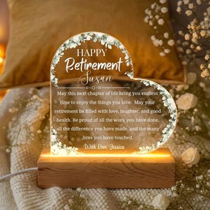 May include: A heart-shaped acrylic retirement gift with the words "Happy Retirement Susan" and a personalized message. The clear acrylic is illuminated by a warm light from a wooden base, and is decorated with floral accents.