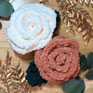 Puede incluir: Dos flores de ganchillo hechas de hilo suave y afelpado. Una flor es de color crema y la otra es de un naranja terracota apagado. Cada flor presenta hojas de ganchillo de color verde oscuro en la base, dispuestas en un diseño de pétalos circulares.