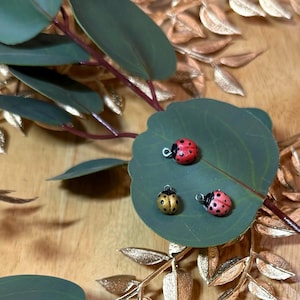 May include: Three small ladybug charms featuring a metal loop at the top for attachment. One charm is bright red with black spots, one is metallic gold with black spots, and one is a lighter pinkish-red with black spots, all resting on a green leaf.