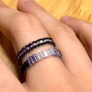 Op de afbeelding: Twee kralenringen met een geweven patroon van kleine, cilindrische kralen. De bovenste ring bestaat uit afwisselend zwarte en doorzichtige witte kralen. De onderste ring toont een verloop van zwart naar paars naar wit.