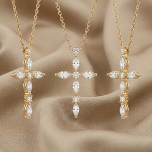 May include: Three gold-colored cross necklaces with clear, sparkling gemstones. Each cross pendant is suspended from a delicate chain. The gemstones are set in a symmetrical pattern, creating a classic and elegant design. The background is a neutral, light brown color.