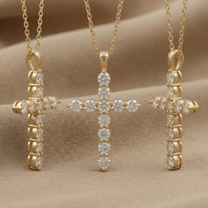 May include: Three gold-colored cross pendants with clear, round gemstones. Each cross hangs from a delicate gold chain. The crosses are displayed against a neutral, tan background, highlighting the jewelry's sparkle.