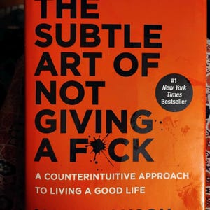 May include: A book with an orange cover and the title "The Subtle Art of Not Giving a F*ck" in large black letters. It includes a black ink blotch graphic and a "#1 New York Times Bestseller" sticker.
