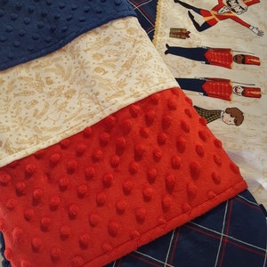 May include: A multi-colored baby blanket with a blue, white, and red patterned design. The blanket is made of soft, textured fabric and features a festive nutcracker design.