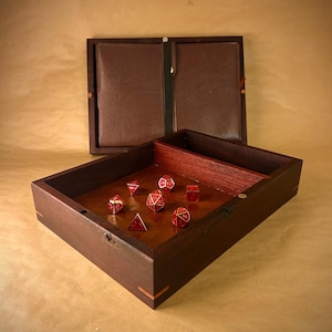May include: A wooden dice rolling tray with a hinged lid, open to reveal a brown leather interior. The tray contains several red dice with gold accents. The tray is made of dark wood with a lighter wood accent.