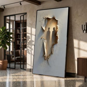 May include: Large framed artwork featuring a textured white surface with a torn section revealing two silhouetted figures. The torn area has a warm, golden hue, creating a contrast with the white background. The artwork is displayed in a modern interior.