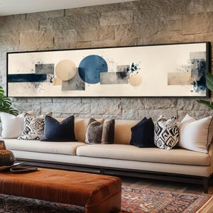 May include: Abstract art print in a black frame. The artwork features geometric shapes in shades of blue, beige, and gray, with a cream-colored background. The print is displayed above a beige sofa with decorative pillows.