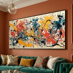 Op de afbeelding: Groot abstract schilderij met een zwarte lijst. Het kunstwerk toont een crèmekleurige achtergrond met spatten en druppels rode, gele, teal, witte en zwarte verf. Het hangt boven een groene bank met decoratieve kussens.
