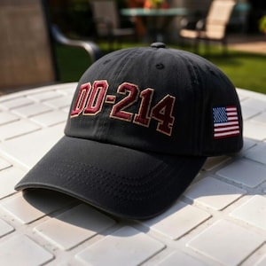 May include: A black baseball cap featuring the embroidered text DD-214 in a maroon font with a gold outline across the front panels. The right side of the cap displays a small, rectangular embroidered patch of the United States flag in red, white, and blue.
