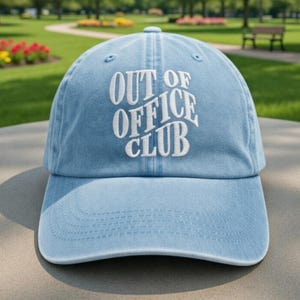 May include: Light blue baseball cap with the embroidered text "OUT OF OFFICE CLUB" in white. The cap has a curved brim and a classic six-panel design. The hat is made of a soft, washed cotton material.