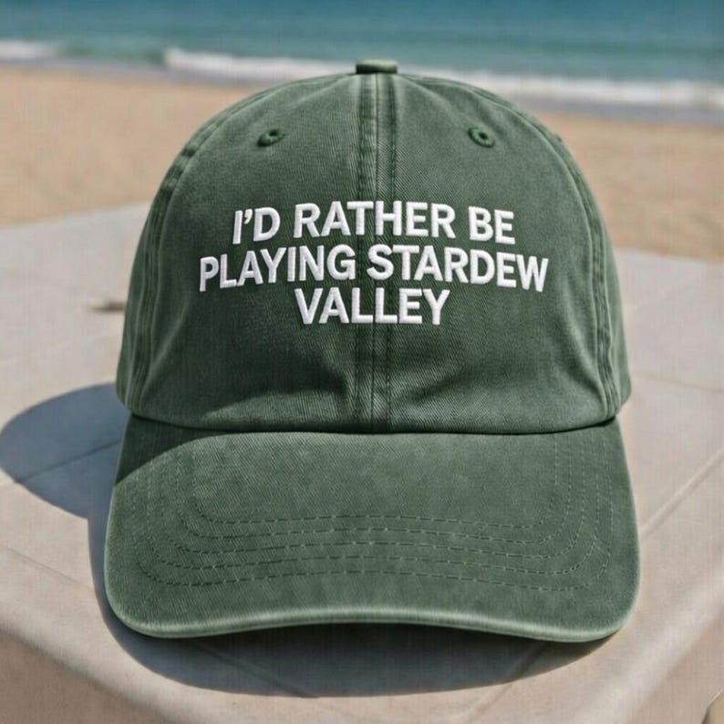 K&ouml;nnte beinhalten: Dunkelgr&uuml;ne Baseballkappe mit dem wei&szlig;en, gestickten Text "I'D RATHER BE PLAYING STARDEW VALLEY". Die Kappe hat einen gebogenen Schirm und einen Stoffriemenverschluss. Der Hut ist aus weichem, abgenutztem Material.