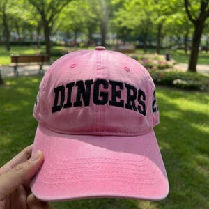 May include: Pink baseball cap with the word "DINGERS" embroidered in black across the front. The cap has a curved brim and a rounded crown. The background is a park with green trees and grass.