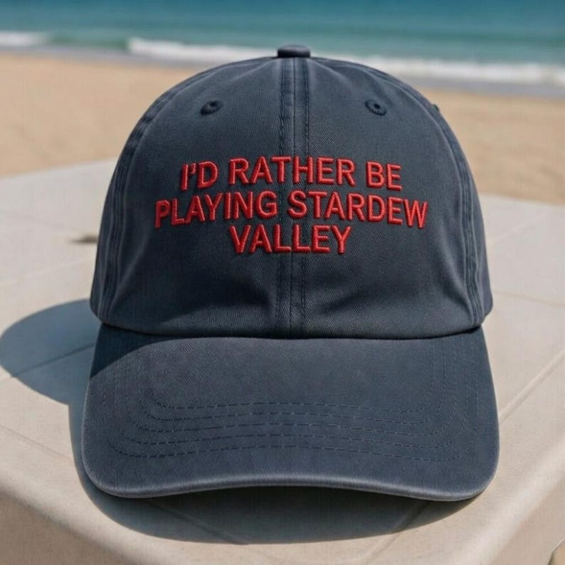 K&ouml;nnte beinhalten: Marineblaue Baseballkappe mit dem rot gestickten Text "I'D RATHER BE PLAYING STARDREW VALLEY". Die Kappe liegt auf einer wei&szlig;en Oberfl&auml;che, mit Strand und Meer im Hintergrund.