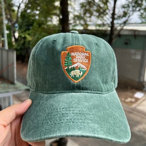 May include: A green baseball cap with a curved brim. The cap features an embroidered patch with the words "NATIONAL PARK SERVICE" and a graphic of a tree, mountain, and bison. The cap is made of a soft, worn material.