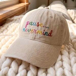 May include: A beige baseball cap featuring the embroidered text "pause... nevermind" in a multi-colored script font. The cap has a structured crown, a curved brim with visible stitching, and eyelets for ventilation on the top panels.