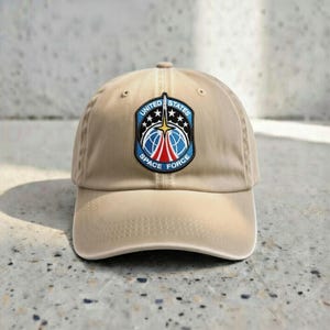May include: A tan baseball cap featuring a patch with the words "UNITED STATES SPACE FORCE" in a blue, red, and white design. The cap has a curved brim and a fabric strap closure.