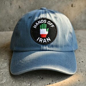 May include: A blue denim baseball cap with a circular patch. The patch features a hand with the colors of the Iranian flag and the words "HANDS OFF IRAN" and "IRAN" in white lettering. The cap is a classic six-panel design.