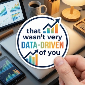 May include: A round sticker with the text "that wasn't very DATA-DRIVEN of you" in a blue and white design. The sticker features a graph and arrow design. The sticker is held over a laptop and other office items.