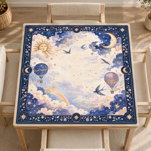 Può includere: Una tovaglia quadrata con un design celeste raffigurante un sole, una luna crescente, mongolfiere e rondini tra nuvole e stelle. Il bordo è blu scuro con accenti floreali e motivi a stella dorati su uno sfondo color crema e blu.
