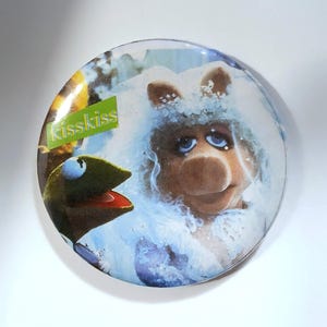 Op de afbeelding: Een ronde knoop met Kermit de Kikker en Miss Piggy van The Muppets. Miss Piggy draagt een witte sluier. De knoop heeft de tekst "Kisskiss" in groen en geel.