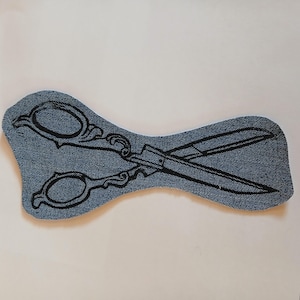 Pode incluir: Um emblema de tecido jeans em forma de tesoura. O emblema apresenta um contorno bordado em preto escuro de uma tesoura ornamentada de estilo vintage, com cabos detalhados e lâminas longas e pontiagudas sobre o material jeans azul.