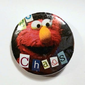 Op de afbeelding: Een ronde knoop met een rood, harig personage met een grote oranje neus en witte ogen. Het woord "Chaos" is in kleurrijke, uitgesneden letters onderaan geschreven. De achtergrond is zwart.
