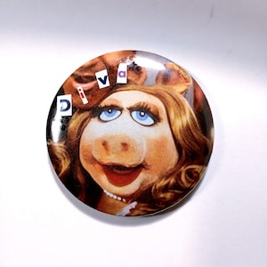 May include: A round button featuring Miss Piggy from The Muppets. The button has a portrait of Miss Piggy with blue eyes, blonde hair, and a pearl necklace. The word "DIVA" is printed on the button.